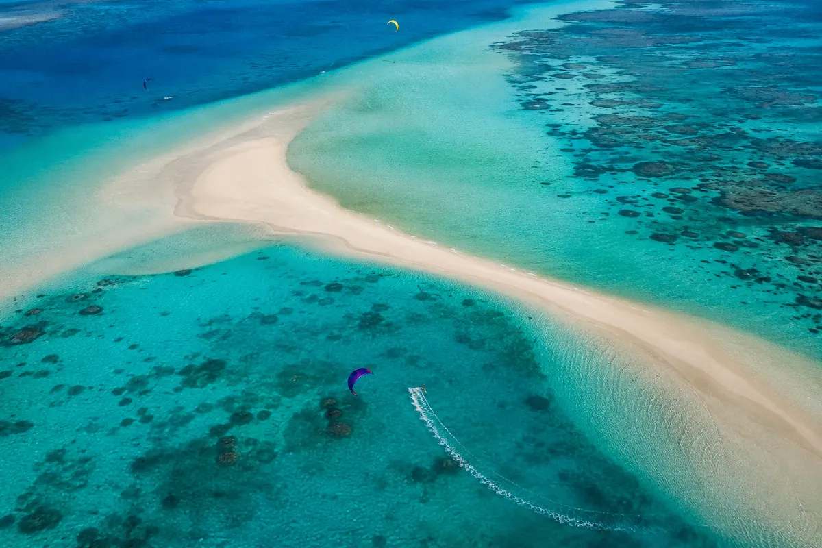 Kuşbakışı çekilen bu büyüleyici manzarada, bembeyaz kum şeritlerinin iki yanında uzanan berrak turkuaz sular üzerinde, rengarenk uçurtmalarıyla sörf yapan birkaç kitesurfçü görülüyor.