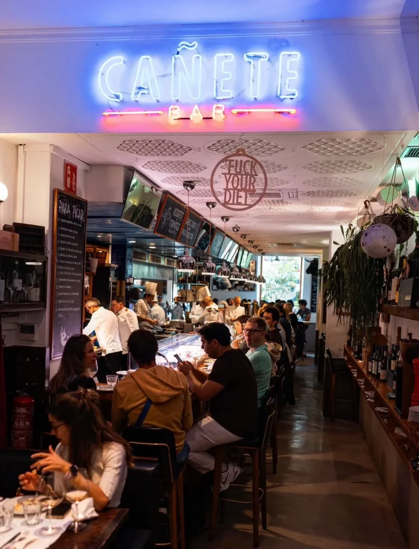 Cañete Bar'ın iç mekanı, parlak mavi neon tabelası altında, kalabalık müşterilerin bar tezgahında ve masalarda oturarak yemek yiyip sosyalleştiği hareketli bir sahneyi gösteriyor; arka plandaki açık mutfakta beyaz şef üniformalı aşçılar hazırlık yaparken, tavandaki FUCK YOUR DIET yazısı eğlenceli bir detay katıyor.