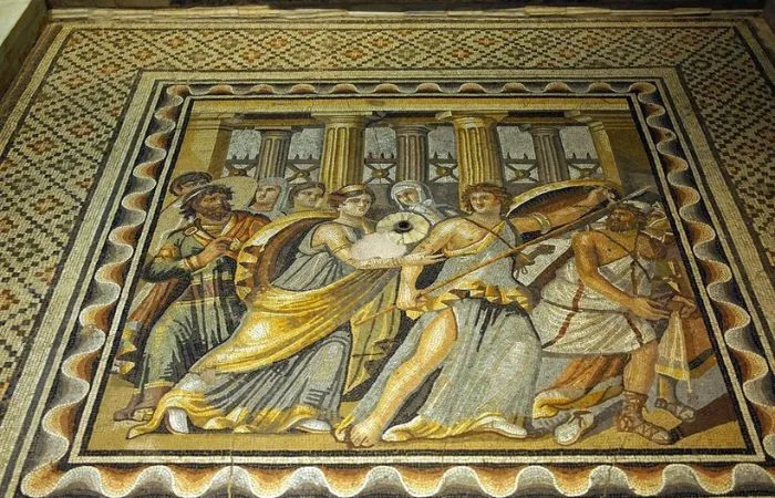 Zeugma Mozaik Müzesi'nden, Truva Savaşı'na katılımıyla tanınan savaşçı Akhilleus'un kalkan ve mızrağıyla çevresindeki figürlerle dinamik bir etkileşim içinde betimlendiği, arka planda klasik sütunlar bulunan detaylı bir mozaik.