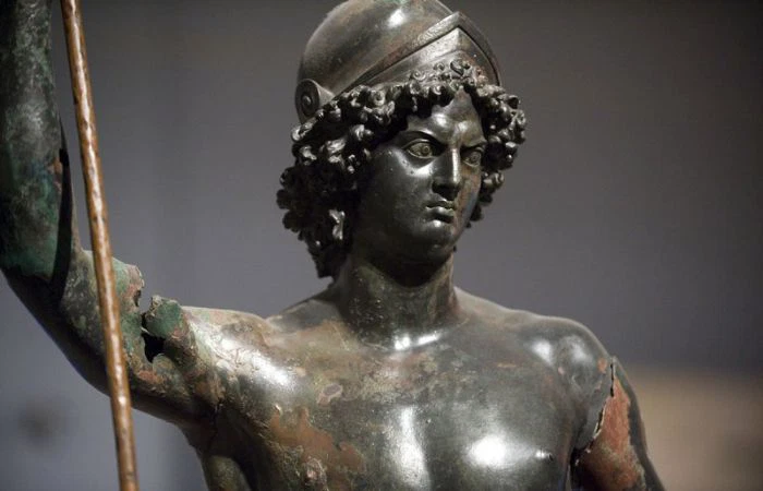 Zeugma Mozaik Müzesi koleksiyonundan öne çıkan Ares (Mars) heykelinin yakından çekilmiş bu görselinde, miğferli, dalgalı saçlı ve kararlı yüz ifadesine sahip, bronzdan yapılmış, yıpranmış ve paslanmış bir tanrı veya savaşçı figürünün göğüs ve baş kısmı, elinde bir mızrak tutarken koyu gri bir fonda yer alıyor.