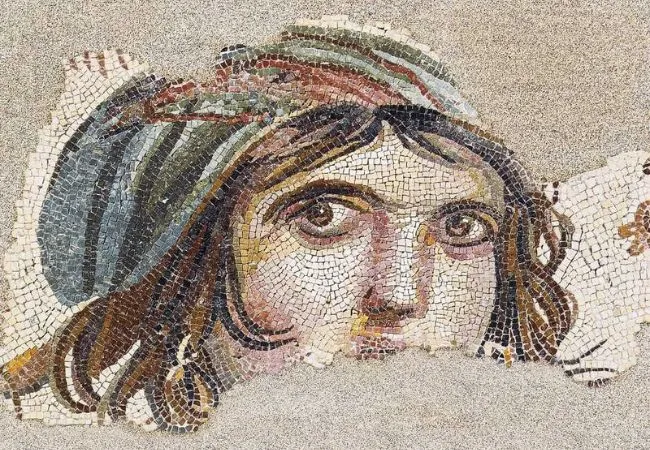 Zeugma Mozaik Müzesi'nin öne çıkan eserlerinden, derin ve etkileyici bakışlarıyla tanınan, kahverengi dalgalı saçlı ve başında renkli bir örtü bulunan genç bir kadının yüzünü gösteren, kısmen yıpranmış antik Çingene Kızı mozaiği detayı.