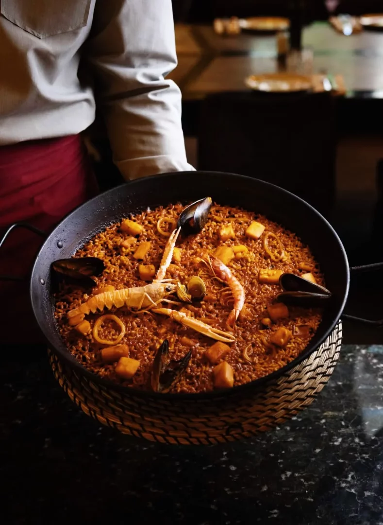 Beyaz gömlekli ve kırmızı önlüklü bir garsonun tuttuğu büyük siyah bir paella tavasında, bol miktarda karides, midye, kalamar halkaları ve belirgin bir langust ile zenginleştirilmiş kızıl-kahverengi pirinçlerden oluşan, iştah açıcı ve rengarenk deniz ürünleri paellası, hasır bir altlık üzerinde gururla sunuluyor.