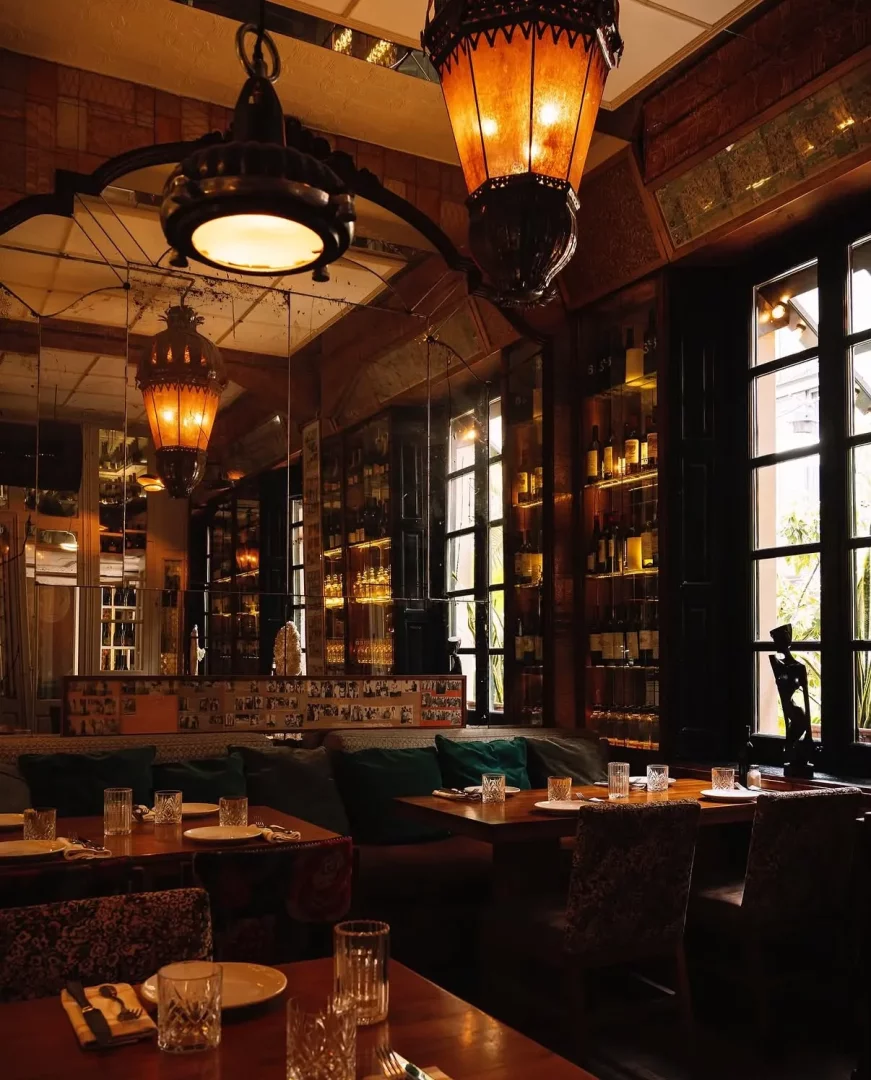 Boca Grande restoranının sıcak ve davetkar atmosferini yansıtan bu geniş iç mekanda, ahşap masalar, koyu yeşil yastıklı oturma alanları, zarif asma lambalar ve şişelerle dolu aydınlık raflar öne çıkıyor, detaylı dekorasyonuyla lüks bir yemek deneyimi sunuyor.