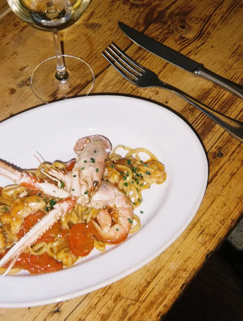 Rustik ahşap bir masa üzerinde, içinde taze deniz mahsulleri (langostinos veya karides), domatesli sos ve yeşil otlarla zenginleştirilmiş linguine tipi makarna bulunan beyaz bir tabak, yanında bir kadeh beyaz şarap, çatal ve bıçak zarifçe yer alıyor.