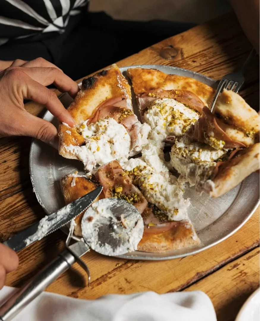 Ahşap dokulu bir masanın üzerindeki metal tepside duran, kenarları hafifçe yanmış, üzeri bol kremalı burrata peyniri, ince dilimlenmiş jambon ve Antep fıstığı parçacıklarıyla dolu gurme pizzadan bir dilimin elle alınışını ve yanında peynir kalıntılarıyla duran bir bıçak ve pizza kesiciyi gösteren detaylı bir görüntü.