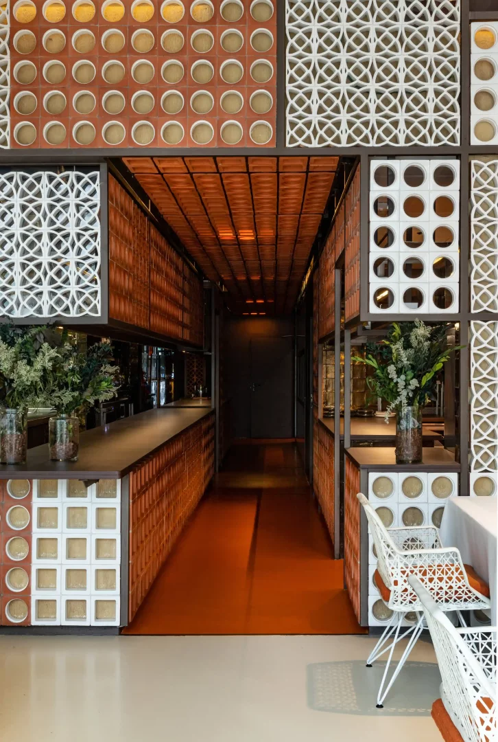 Kırmızı tuğla ve beyaz geometrik desenli panellerle süslü, sıcak turuncu zeminli uzun bir koridor ve yemek alanının görüldüğü bu modern restoran tasarımı, bitki aranjmanları ve beyaz sandalyelerle özgün bir atmosfere sahip.