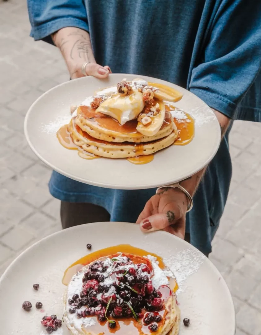 Mavi bir gömlek giyen ve dövmeleri olan bir kişinin ellerinde, üstte muz dilimleri, çırpılmış krema ve fındıklarla süslenmiş karamel soslu üç kat pancake, altta ise böğürtlen ve frambuaz gibi çeşitli meyvelerle zenginleştirilmiş, pudra şekeri serpilmiş bir diğer lezzetli pancake tabağı, brunch veya kahvaltı keyfi için sunuluyor.