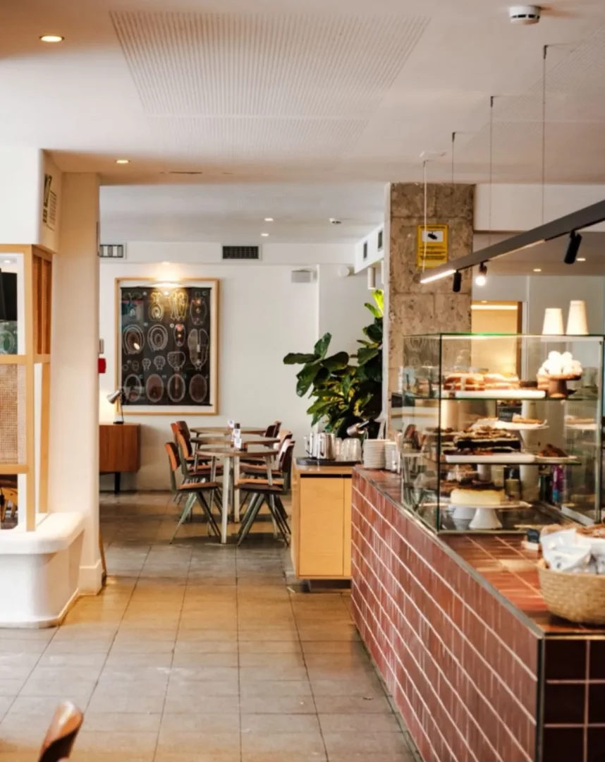 Modern ve davetkar bir kafe iç mekanında, ön planda kırmızımsı kahverengi fayanslarla kaplı uzun tezgahın arkasında cam vitrinde çeşitli kekler ve hamur işleri sergilenirken, arka planda ahşap masalar ve sandalyelerle düzenlenmiş oturma alanı ile duvarda bilimsel illüstrasyonlar içeren büyük bir tablo görülmektedir.
