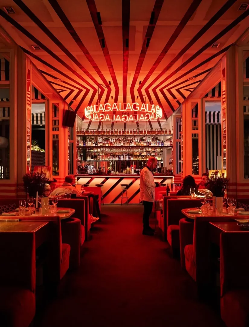 Kırmızı ışıklarla dolu, modern tasarımlı bu lüks restoranda, tavandaki çarpıcı siyah-kırmızı çizgili desenler ve barın üzerindeki parıldayan GALA neon tabelası, atmosferik bir gala gecesi veya özel etkinlik için şık bir ortam sunarken, kırmızı kadife koltuklarda oturan misafirler masalarda sosyalleşiyor.
