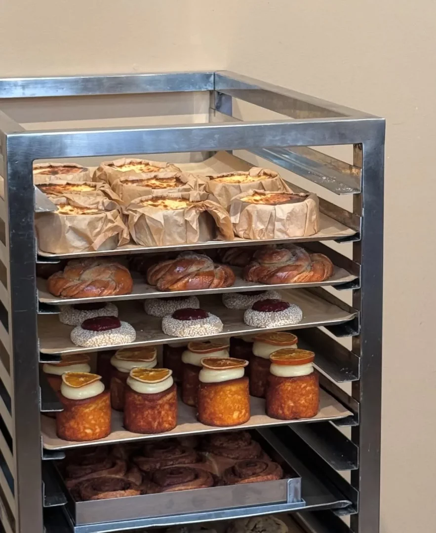 Metal bir raf üzerinde, kahverengi kağıtlara sarılı peynirli tartlar, örgülü çörekler, beyaz şekerli kaplamalı ve kırmızı reçelli küçük tatlılar, krema ve kurutulmuş portakal dilimli silindirik kekler ile tarçınlı rulolar gibi çeşitli taze fırın ürünleri sergileniyor.