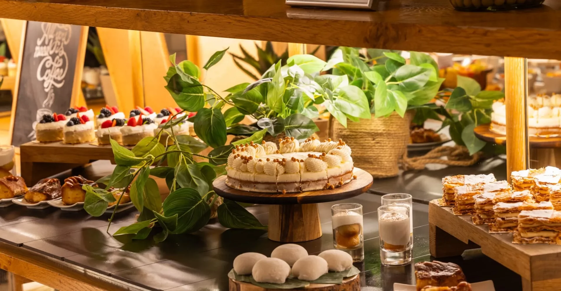 Bir otel veya restoranda özenle hazırlanmış, ortasında dilimlenmiş kremalı ve topçuk süslemeli büyük bir pastanın durduğu, yanında karışık meyveli tartletler, kat kat pudra şekerli milföy tatlıları ve yeşil bitkilerle dekore edilmiş zengin bir tatlı büfesi sunumu.