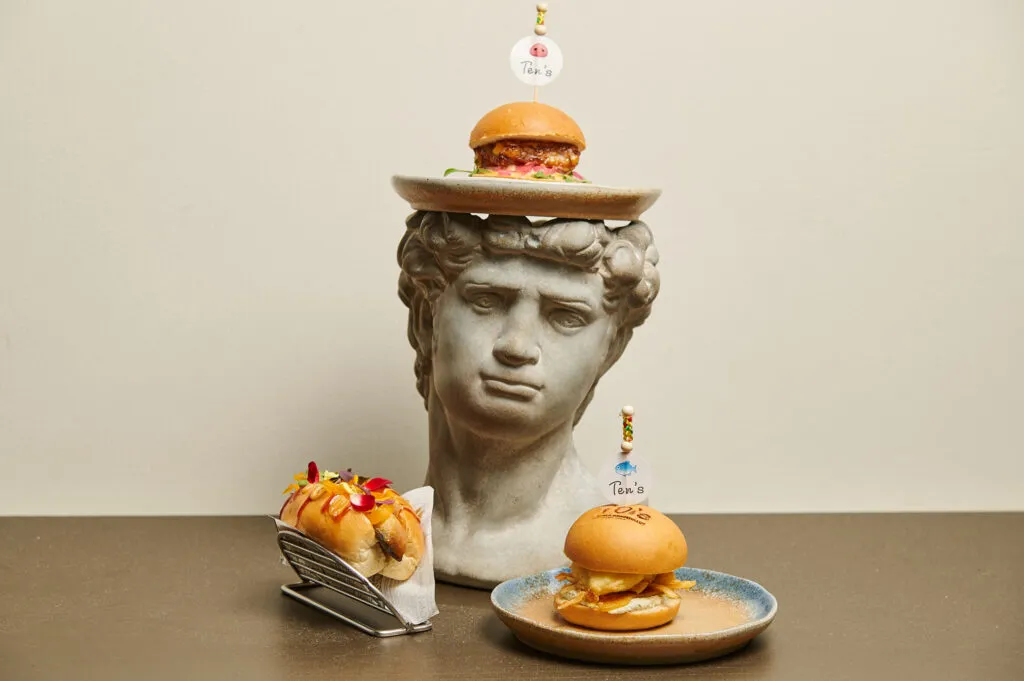 Gri renkli David büstünün üzerinde bir tabakta duran küçük bir burger ile solunda metal standda rengarenk çiçeklerle süslenmiş sandviç ve sağında mavi kenarlı bir tabakta servis edilen patates kızartmalı burger, Ten's markasının sanatsal ve dikkat çekici yiyecek sunumunu gözler önüne seriyor.