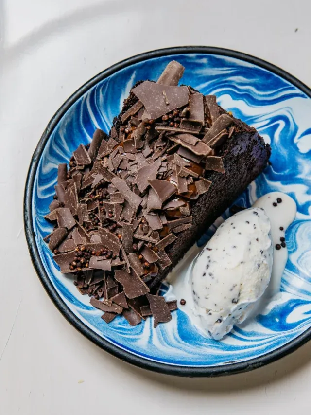 Koyu çikolatalı kek veya brownie dilimi, üzerine serpilmiş bol miktarda ince çikolata parçacıkları ve çıtır çikolata topları ile zenginleştirilmiş, yanında benekli beyaz dondurma topuyla, mavi ve beyaz mermer desenli şık bir tatlı tabağında sunuluyor.