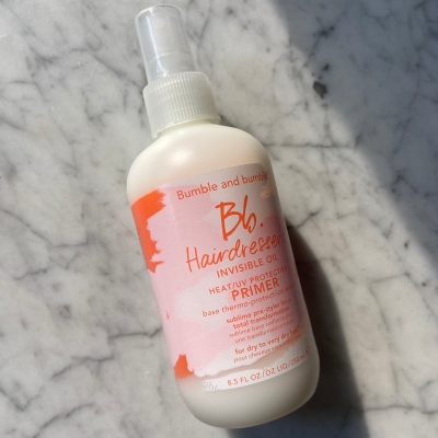 Mermer bir yüzeyde duran, pembe ve turuncu etiketli, ısı ve UV koruması sağlayan beyaz renkli Bumble and Bumble Hairdresser's Invisible Oil Heat/UV Protective Primer sprey şişesi, boyalı saçlar için güneş ışınlarına ve ısıyla şekillendirmeye karşı etkili bir bakım önerisi sunuyor.