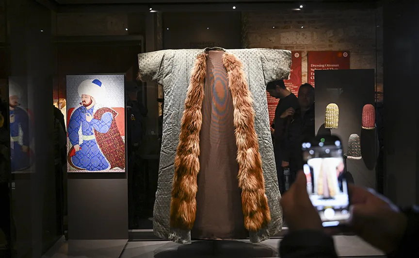 Topkapı Sarayı'nda sergilenen, açık renkli, desenli, uzun ve önü kahverengi kürklü bir padişah kaftanı, yanında mavi giysili bir padişah portresi ve küçük başlıklarla birlikte ziyaretçilerin cep telefonlarıyla fotoğraf çektiği bir müze vitrininde.