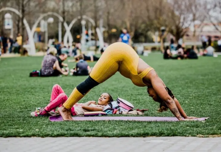 Yeşil çimler üzerinde mor bir yoga matında aşağı bakan köpek pozisyonunda esneyen koyu tenli bir kadın, yanında rahatça yatan küçük bir kız çocuğu ile anne adaylarının doğuma hazırlık sürecinde fiziksel aktivitenin önemini vurguluyor.