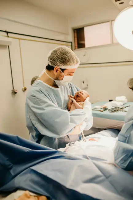 Maskeli bir sağlık profesyoneli, ameliyathanede yeni doğmuş, göbek bağı hâlâ bağlı ve yüksek sesle ağlayan bir bebeği nazikçe tutarken görülüyor; bu an, anne adayları için sağlıklı bir doğum sürecinin ve doğuma hazırlığın önemini vurguluyor.