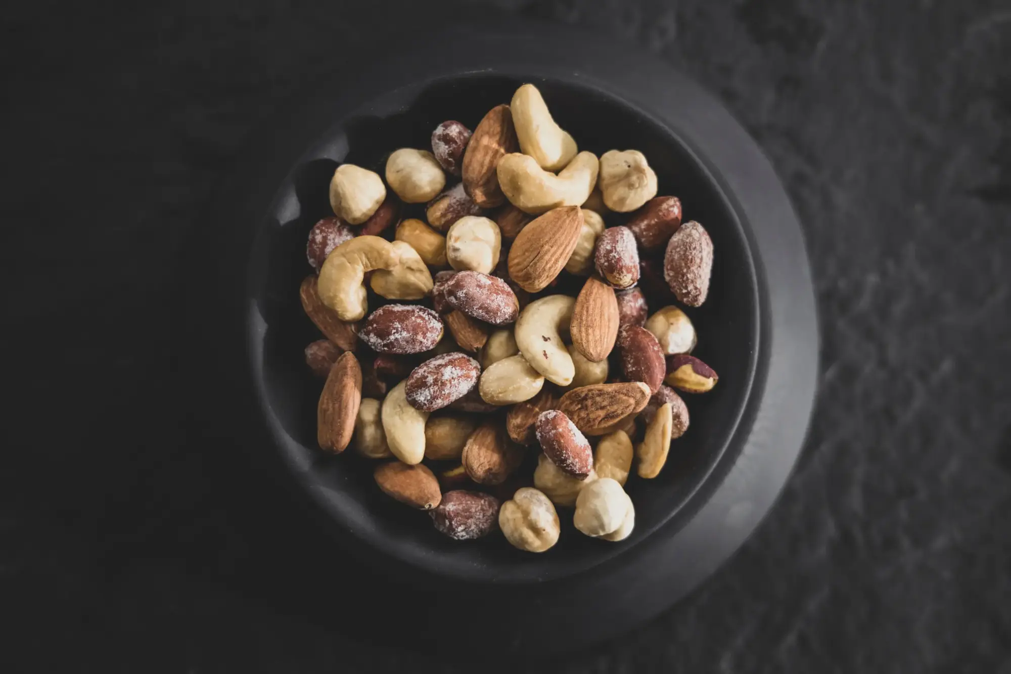 Bir kase içinde karışık kuruyemişler; yer fıstığı, badem ve kaju gibi besin alerjisine yol açabilecek yaygın protein kaynakları, koyu renkli bir zeminde yukarıdan çekilmiş bir açıyla görülüyor.