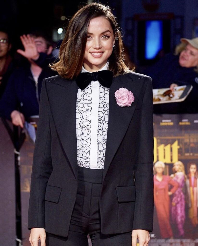 Ana de Armas, Londra Film Festivali'nde sergilediği ikonik maskülen stiline uygun olarak, geniş omuzlu siyah takım elbisesi, fırfırlı beyaz gömleği, kadife papyonu ve yakasındaki zarif pembe karanfiliyle kameraya gülümseyerek poz veriyor.