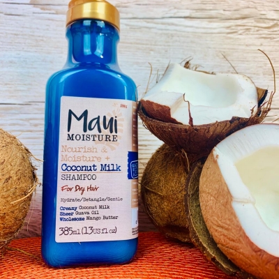Mavi renkli, altın kapaklı bir Maui Moisture Coconut Milk sülfatsız şampuan şişesi, etiketinde kuru saçlar için özel formülü ve besleyici içeriği belirtilerek, parçalanmış ve bütün hindistan cevizi kabukları arasında ahşap bir yüzeyde yer alıyor, saçları nazikçe temizleyip nemlendirme konusundaki vaadini vurguluyor.
