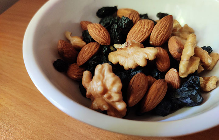 Ahşap bir masa üzerinde, parlak beyaz bir kase içerisinde, saç derisi sağlığını destekleyen ve Omega-3 gibi faydalı yağlar içeren badem, ceviz ve koyu renkli kuru üzüm tanelerinden oluşan besleyici bir karışım yakın planda yer alıyor.
