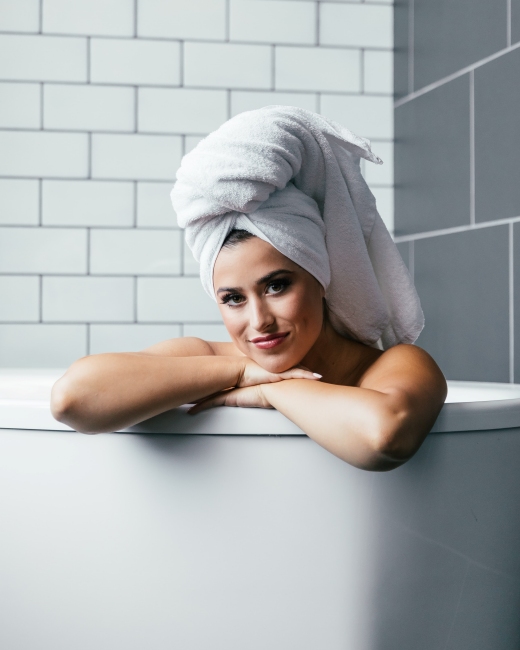 Kadın, banyo sonrası saç bakımı rutinini uygulayacak gibi, saçlarını beyaz havluyla sarmış ve küvetin kenarına yaslanarak gülümsüyor, sağlıklı, parlak ve güçlü saçlar için saç sirkesi ve sirkeli saç toniklerinin faydalarını düşünürken.