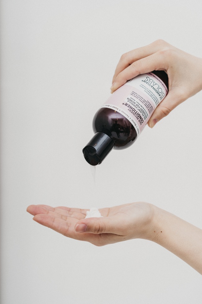 Kadın elleri açık renk bir arka plan önünde, bir elinde SCANDINAVIAN BIOLABS Hair Recovery Formula Conditioner+ yazılı şişeden diğer elin avucuna beyaz bir saç kremi dökerken, bu görsel modern saç bakımı ürünleri ve rutinlerinin önemini vurguluyor.