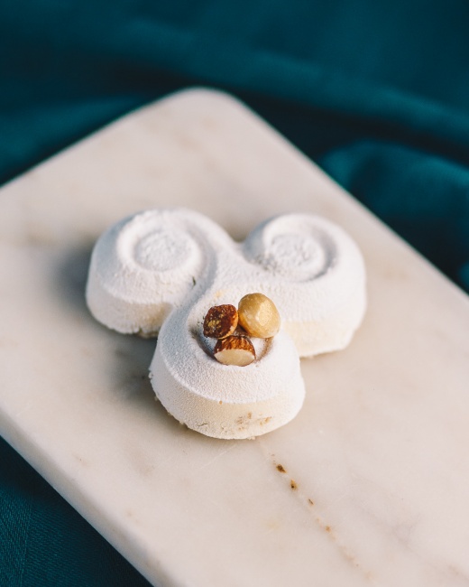 İzmir'in modern pastacılık anlayışını yansıtan La Floran Patisserie'den, beyaz kadifemsi dokulu, kıvrımlı ve zarif şekilli bir tatlı, ortasındaki minik fındık taneleriyle mermer sunum tabağında koyu teal bir zemin üzerinde çekici bir şekilde sergileniyor.