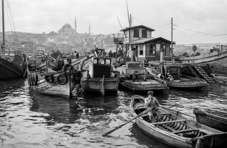 Ara Güler'in kadrajından, İstanbul'un eski dönemlerine ait canlı bir liman hayatını betimleyen siyah beyaz fotoğraf; çok sayıda ahşap teknenin yanaştığı hareketli iskelede, önde bir kayıkta kürek çeken bir figür ve uzakta yükselen Süleymaniye Camii'nin etkileyici silüeti görülüyor.