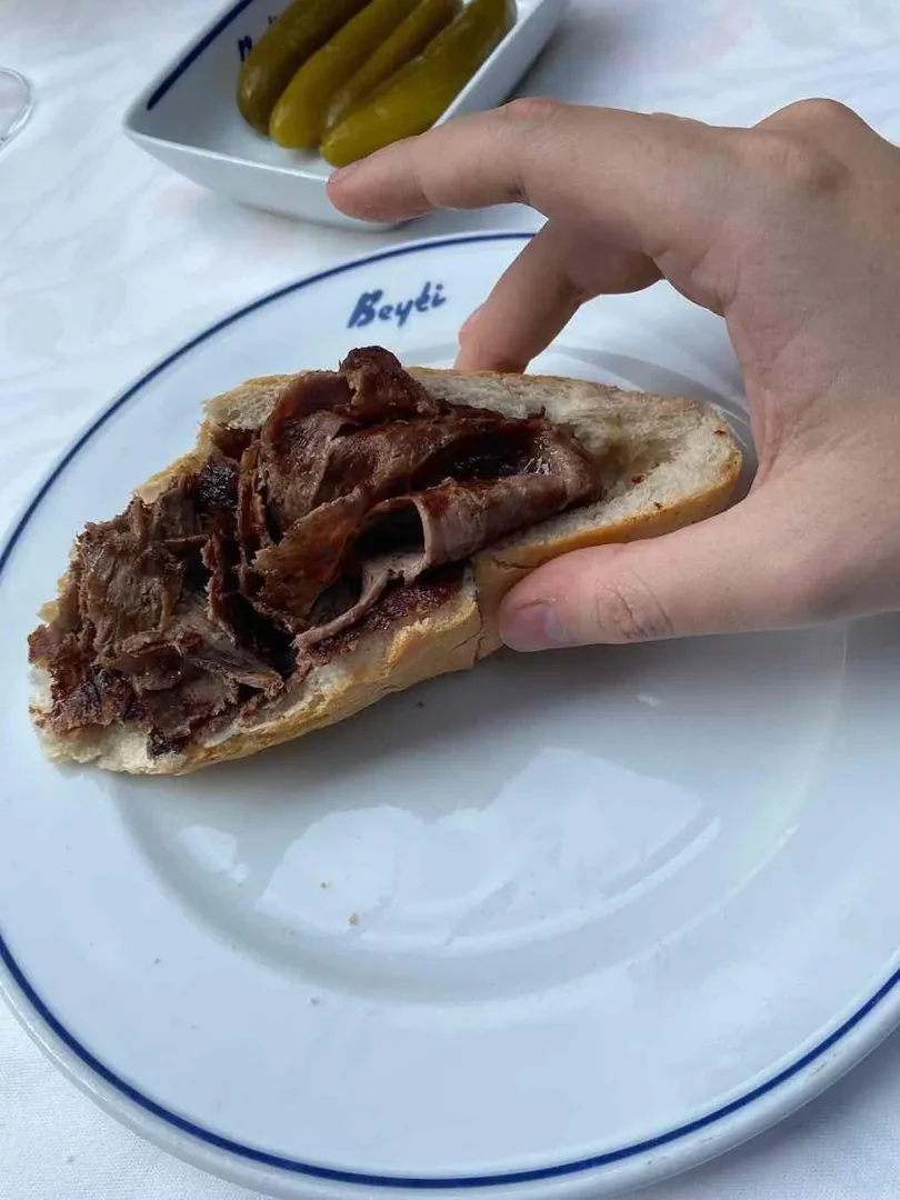 Bir elin, Beyti restoranının mavi kenarlı beyaz tabağında duran, bol etli ekmek arası döneri tuttuğu ve arka planda salatalık turşularının belirgin şekilde görüldüğü, Türk mutfağının lezzetlerini yansıtan iştah açıcı bir an.