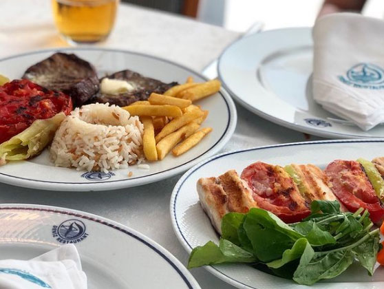 İzmir Deniz Restoran logosu taşıyan şık servis tabaklarında, nefis bir balık ziyafeti sunumu göze çarpıyor; tereyağlı pilav, çıtır patates kızartması, közlenmiş domates ve biber ile tamamlanmış ızgara balık filetoları, arka planda hafifçe görünen içecekle ve ön plandaki ızgara balık şişleri ve taze roka ile birlikte İzmir'in en iyi restoranlarından birinin özenli menüsünü yansıtıyor.