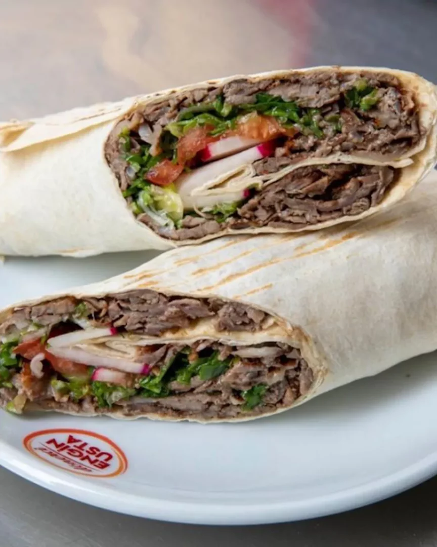 Beyaz bir tabağa yarısı kesilmiş iki adet et döner dürüm sergileniyor; lavaş ekmeği içinde bolca döner eti, taze yeşillikler, domates ve turp dilimleri net bir şekilde görülürken, tabağın köşesinde Engin Usta logosu dikkat çekiyor.