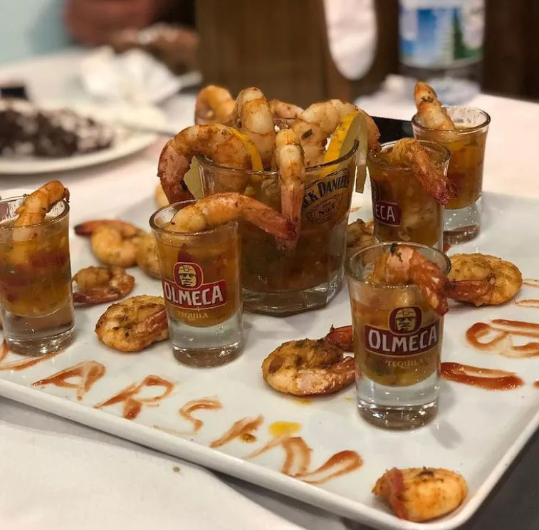 Beyaz dikdörtgen bir servis tabağında, içlerinde Jack Daniel's ve Olmeca Tequila marka shot bardakları da bulunan bardaklara ve tabağa özenle yerleştirilmiş baharatlı pişmiş karidesler, tabağın üzerinde sosla yazılmış ARSAN kelimesiyle Foça'daki bir restoranın dikkat çekici deniz ürünleri sunumunu yansıtıyor.