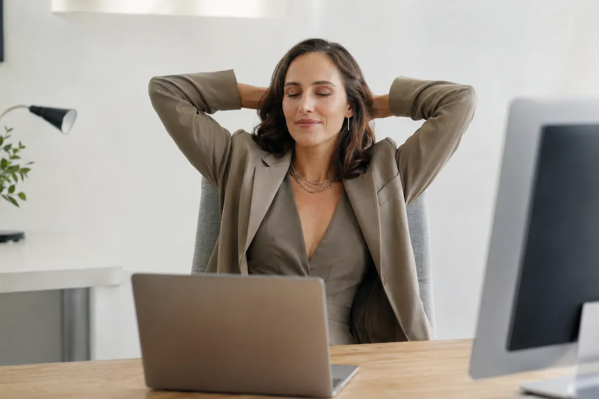 İş dönüşü çalışma motivasyonunu artırmak için molaların ne kadar kritik olduğunu gösteren bu görselde, bir kadın modern ofis sandalyesinde rahatlamış bir şekilde oturarak gözleri kapalı, ellerini başının arkasında birleştirmiş, dinlenirken verimli bir çalışma günü için enerji topluyor.