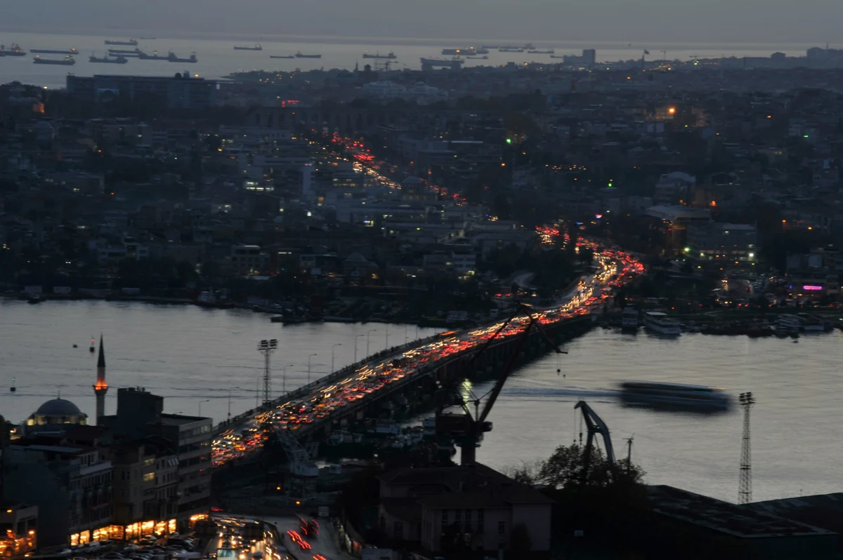 Alacakaranlıkta İstanbul'un ışıklarla parlayan şehir manzarasında, Haliç üzerindeki köprüde ve bağlantı yollarında oluşan yoğun İstanbul trafiği, uzun kırmızı ve beyaz araç ışık çizgileriyle adeta bir enerji akışını andırırken, bu kentsel hareketliliğin karbon ayak izine etkisini düşündürüyor. Uzakta denizde bekleyen gemiler ve belirgin bir minare de kadrajda.