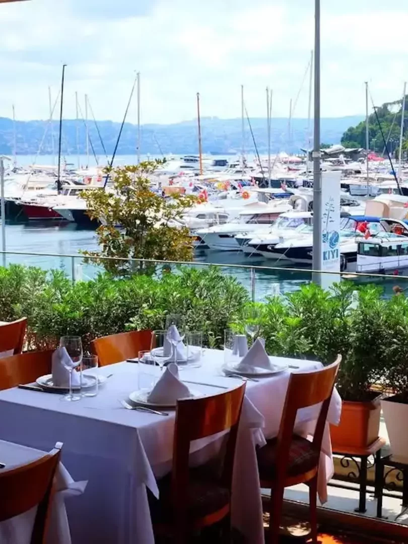 İstanbul'daki Kıyı Restaurant'ın ferah terasından çekilen bu görselde, beyaz masa örtüleriyle özenle hazırlanmış masalar ön planda yer alırken, arka planda Boğaz'ın sakin sularında demirlemiş yelkenli ve yatlarla dolu bir marina ile karşı kıyıdaki yeşil tepeler eşsiz bir manzara sunuyor.