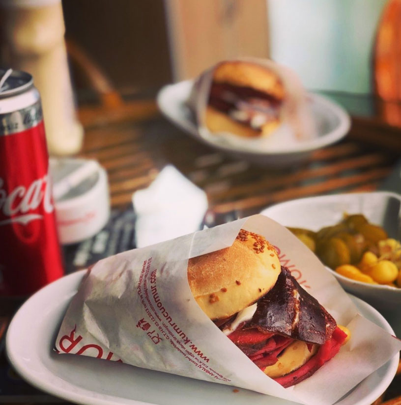 İzmir'in en iyi sokak lezzetlerinden Kumrucu Ömür'e ait, susamlı ekmeği, sucuk, kaşar ve pastırma ile bolca doldurulmuş, yanında soğuk Coca-Cola ve turşuyla sunulan iştah açıcı bir kumru sandviçi.