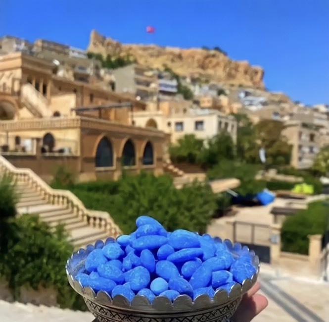Mardin'in eşsiz taş mimarisine sahip tarihi binalarının ve dağlık manzarasının önünde, elde tutulan geleneksel bir metal kapta duran parlak mavi badem şekerleri, şehrin ünlü yerel lezzetlerinden ve keyifli hediyeliklerinden birini gözler önüne seriyor.
