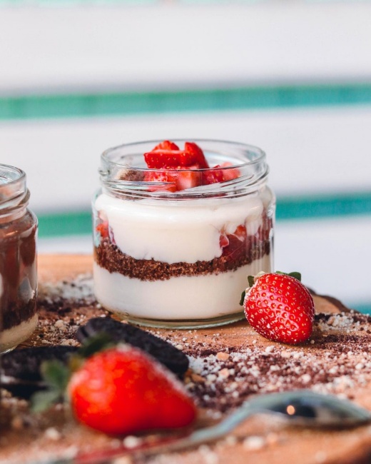 İzmir'in en iyi pastanelerinden biri olan Mint Patisserie'de sunulan, ahşap bir sunum tahtası üzerinde yer alan, tabanı bisküvi kırıntıları, ortası beyaz krema ve üzeri bolca küp doğranmış taze çileklerle dolu, Magnolia tarzı cazip bir tatlı net bir şekilde görülüyor, etrafında taze çilekler ve bisküvi parçacıkları dağınık halde bulunuyor.