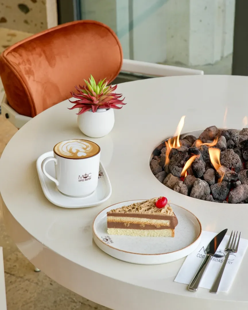 MOC Coffee Roastery'nin modern ve sıcak atmosferinde, beyaz yuvarlak bir masanın üzerinde, latte artlı fincan kahve, kiraz süslemeli kremalı bir dilim pasta, şık bir bitki ve masanın ortasındaki yanan gazlı şömine ile davetkar bir kafe sahnesi görülüyor.