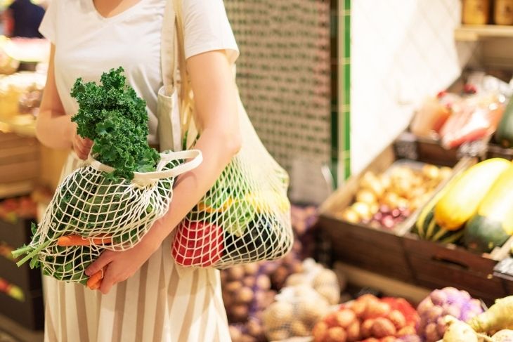 Market alışverişi yapan bir kişi, sürdürülebilir yaşam felsefesine uygun olarak plastik yerine tekrar kullanılabilir file çantalarla taze yeşillikler, havuç, kırmızı biber ve patlıcan gibi ürünleri seçerken görülüyor, bu da mutfakta sıfır atık hedefine katkıda bulunuyor.