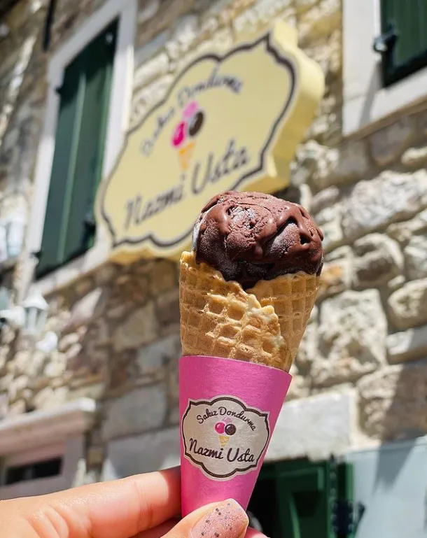 Pembe ambalajlı waffle külahında, hafifçe eriyerek aşağı doğru akan tek top koyu renk çikolatalı dondurmayı tutan ojeli bir el görünüyor; arka planda bulanık şekilde bir taş bina ve Sakız Dondurma Nazmi Usta yazılı sarı dükkan tabelası seçilirken, bu kare Nazmi Usta'nın meşhur dondurmalarından birini tadarken yaz gününün keyfini çıkaran bir anı yakalıyor.