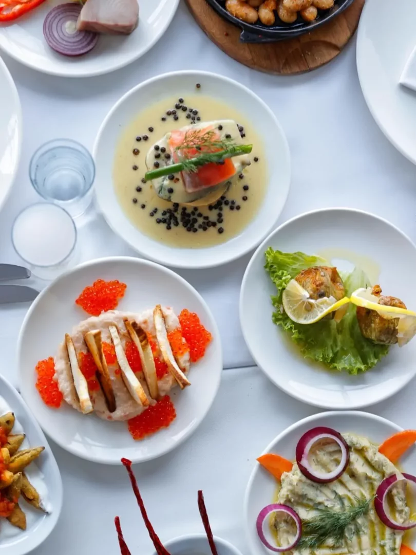 Beyaz masa örtüsüyle kaplı bir masada, ortasında kremalı soslu somon ve karabiberli bir meze, solunda parlak turuncu havyarlı balık ezmesi, sağında limon ve yeşilliklerle servis edilen ızgara balık lokmaları ve kırmızı soğan halkalarıyla süslenmiş dip soslar gibi, çeşitli lezzetli balık ve deniz ürünleri mezeleriyle donatılmış zengin bir sofra.