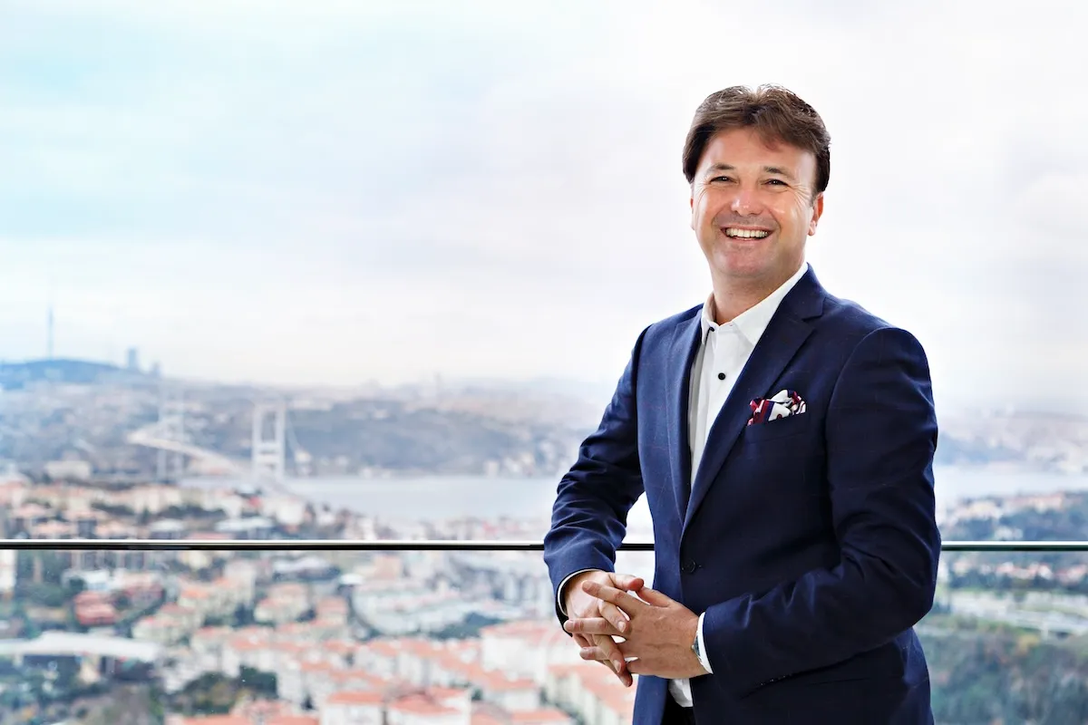 Tarihçi, yazar ve TV programcısı Saffet Emre Tonguç, lacivert bir takım elbise ve beyaz gömleğiyle, ardında siluet halinde görünen Boğaz Köprüsü ve İstanbul manzarasıyla, şehre dönüş temasına uygun olarak samimi bir gülümsemeyle fotoğraf makinesine bakıyor.
