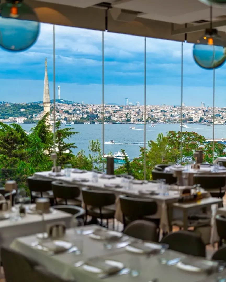 Boğaziçi'ne nazır şık bir restoranda, beyaz örtülü masalar ve koyu renkli sandalyeler ön planda yer alırken, geniş camların ardında teknelerin seyrettiği masmavi Boğaz, tarihi bir kule ve modern kentsel yapılarla çevrili İstanbul silüeti nefes kesici bir manzara sunuyor.
