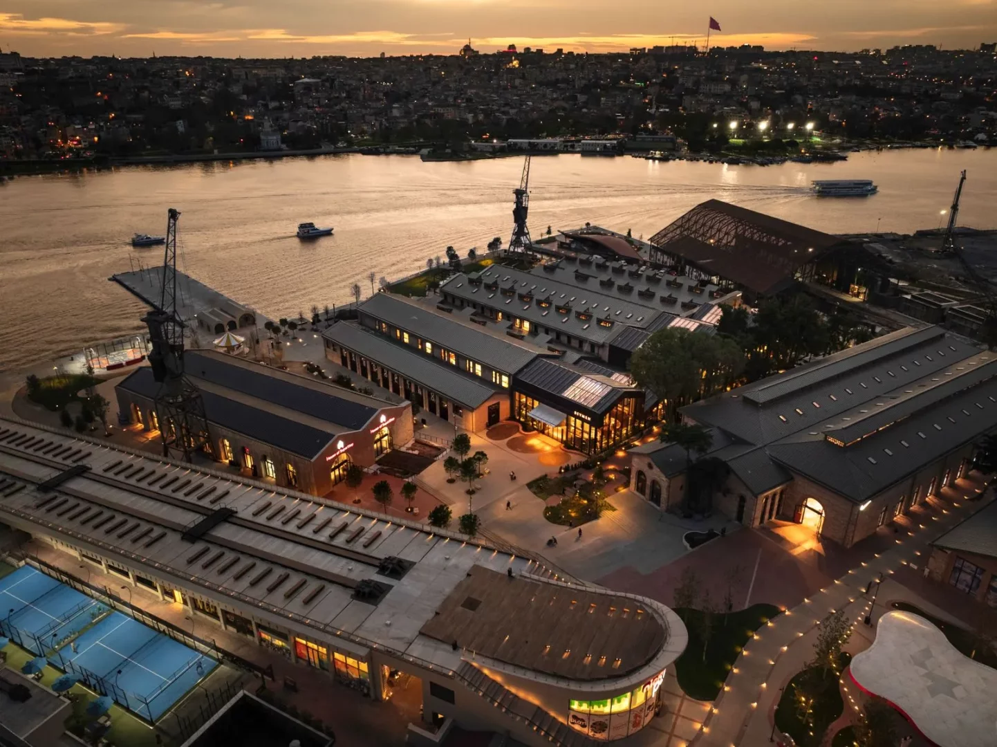Gün batımının turuncu tonlarıyla aydınlanan İstanbul Haliç kıyısındaki Tersane İstanbul'un havadan çekilmiş bu görüntüsünde, eski tersane binaları modern tesisler, ışıklı yürüyüş yolları ve hareketli bir atmosferle birleşerek şehre yeni bir soluk katıyor.