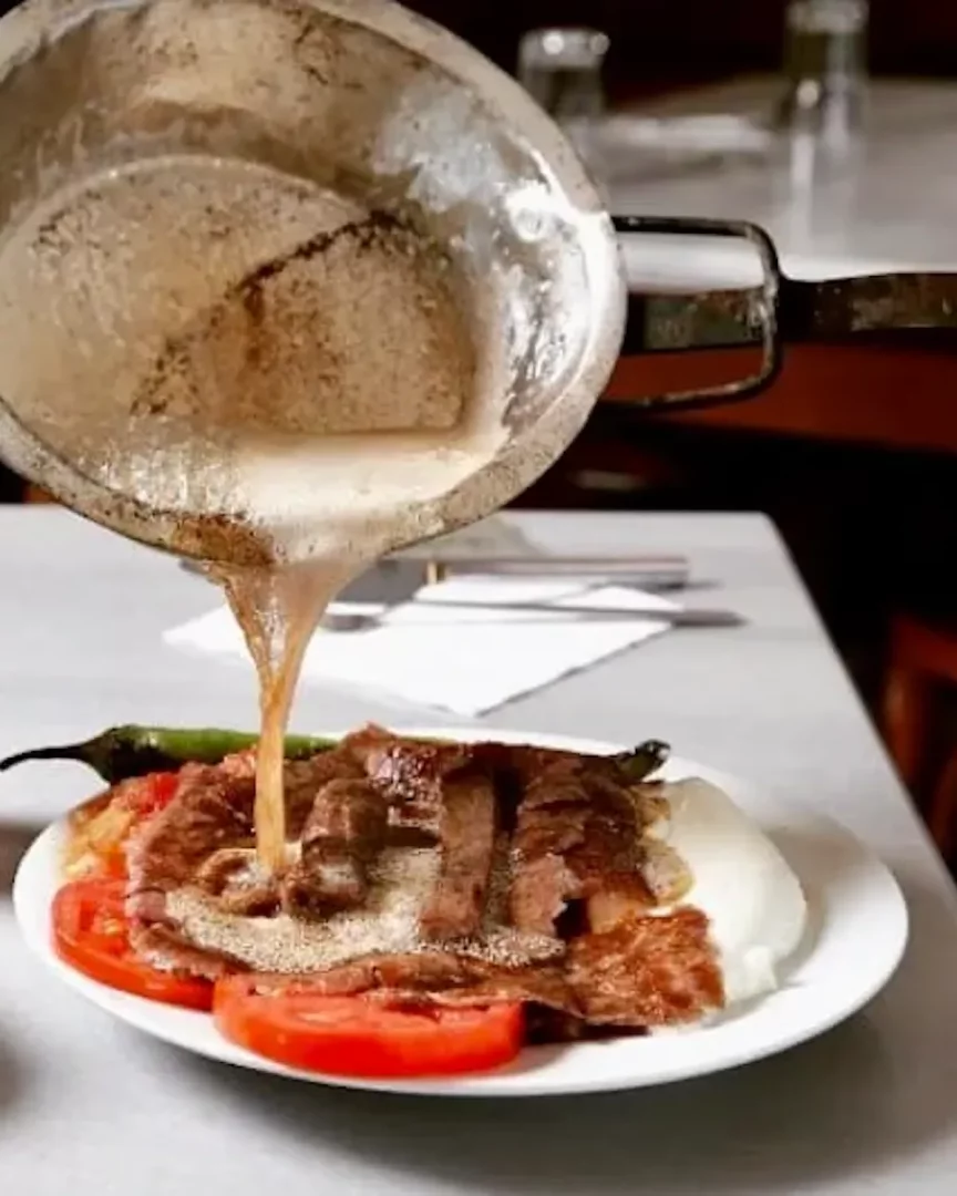 Üzerine sıcak tereyağı gezdirilen nefis bir İskender kebap tabağı, ince kesilmiş et dilimleri, kızarmış pideler, közlenmiş yeşil biber ve domateslerin yanında beyaz yoğurtla servis edilmiş halde, Uludağ Kebapçısı geleneğini yansıtan doyurucu bir lezzet şöleni sunuyor.
