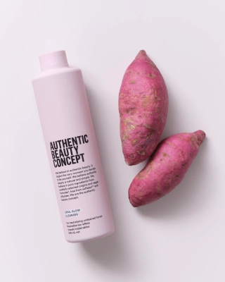 Bir ışık pembe Authentic Beauty Concept Cool Glow Cleanser şampuan şişesi, ürünün mor tatlı patates özleri içerdiğini görsel olarak destekleyen iki taze mor tatlı patates ile beyaz bir zeminde sergileniyor, bu da boyalı saçlar için renk tonunu dengeleyici önemli bir bakım önerisi olarak sunuluyor.