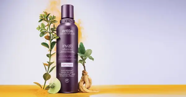 Açık mor ve sarı tonlarında geçişli bir arka plan üzerinde, doğal kök ve yapraklı bitkilerle çevrili, vegan sertifikalı koyu mor Aveda Invati Advanced Activating Şampuan şişesi, saç dökülmesine karşı etkili bir vegan saç bakım ürünü olduğunu vurguluyor.