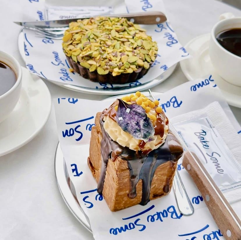Bake Some'dan modern ve yaratıcı sunumlarla; alt kısmı kızarmış ekmek tabanlı kalın bir hamur, üzerinde erimiş çikolata sosu, krema, sarı taneler ve mor bir yaprakla süslenmiş özgün bir tatlı ile yemyeşil bol fıstıkla kaplı yuvarlak bir tartın bulunduğu iki lezzetli porsiyon, İzmir’in en iyi pastaneleri deneyimini yansıtan keyifli bir görüntü oluşturuyor.